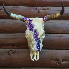 a cow skull with purple and white flowers on it's horns hanging from the side of a log cabin