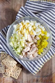 a glass bowl filled with chicken salad next to crackers on a striped towel and wooden table