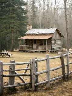 an old log cabin sits in the woods near a fence and picnic table with benches