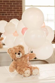 a brown teddy bear sitting on top of a table with white balloons in the air