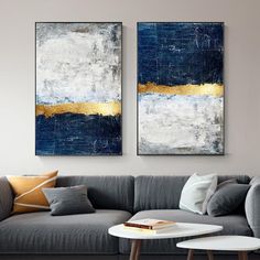 two paintings hang on the wall above a gray couch in a living room with white and gold accents