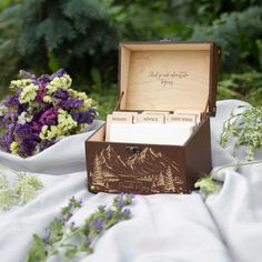 an open wooden box sitting on top of a white cloth covered table next to purple and green flowers