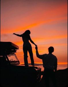 a man holding the hand of a woman standing on top of a car at sunset