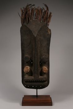 an african mask with two eyes and feathers on it's head, sitting on a wooden stand