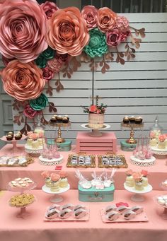 a table topped with lots of cakes and cupcakes covered in frosting next to giant paper flowers