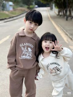 two young children standing next to each other on the side of a road with trees in the background