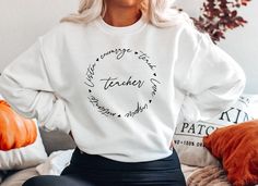 a woman sitting on a bed wearing a white sweatshirt with the words teacher written in black