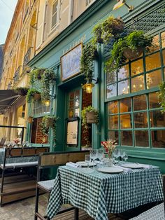 an outdoor dining area with tables and chairs in front of a green building, surrounded by potted plants