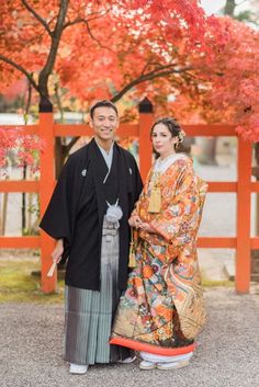 a man and woman dressed in traditional japanese garb standing next to each other under a tree with red leaves