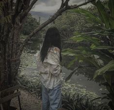 a woman standing next to a tree near a pond and forest with lots of green plants