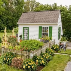 a small white house with green shutters and flowers in the front yard, next to a bike