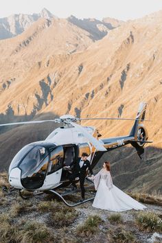 a bride and groom standing in front of a helicopter on top of a mountain with mountains in the background