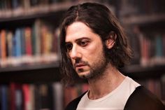 a man with long hair standing in front of bookshelves