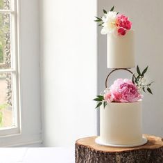 two tiered wedding cake with pink and white peonies on top, sitting in front of a window