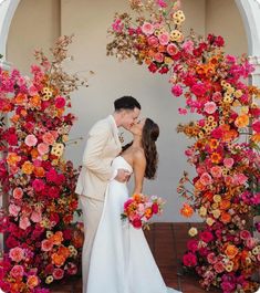 a bride and groom kissing in front of an archway with flowers on the wall behind them