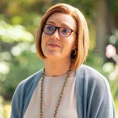 a woman wearing glasses and a necklace looks off to the side with trees in the background