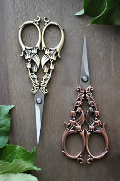 two pairs of scissors sitting next to each other on top of a wooden table with green leaves