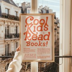 a sign that says cool kids read books hanging on a window sill in front of a cityscape