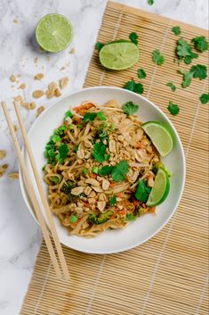 a white bowl filled with noodles and vegetables next to chopsticks on a bamboo mat