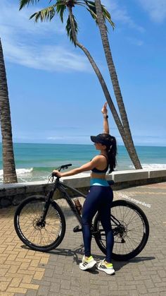 a woman standing next to a bike on the beach with palm trees in the background