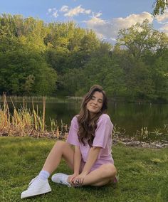 a young woman sitting on the grass next to a lake with trees in the background