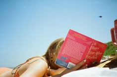 a woman laying down reading a book on the beach with her head in her hands