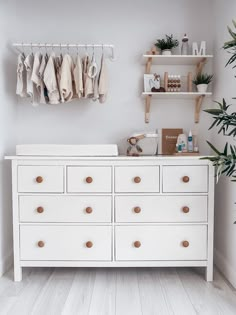 a white dresser with lots of clothes hanging on it's hooks and shelves next to a potted plant