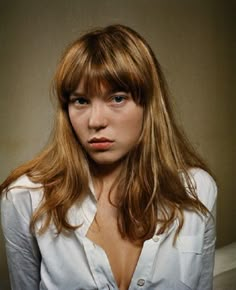 a woman with long hair wearing a white shirt and looking at the camera while standing in front of a wall