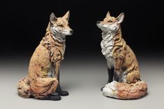 two figurines of foxes sitting next to each other on a gray surface with black background