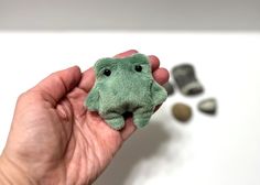 a hand holding a small green stuffed animal in front of some rocks and pebbles on a white surface