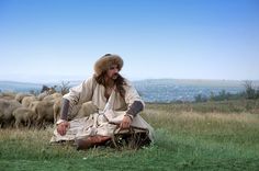 a man with long hair sitting on the ground in front of a herd of sheep