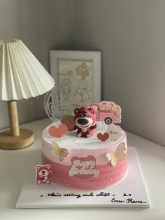 a pink birthday cake with a teddy bear on top and hearts around the edges, sitting on a table next to a lamp