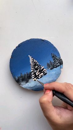 someone is painting a snowy scene on a blue plate with trees in the snow and holding a paintbrush