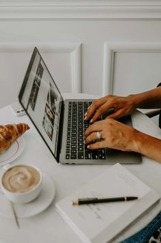 a person sitting at a table using a laptop computer with a croissant in front of them