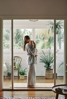 a woman holding a baby standing in front of a sliding glass door with potted plants