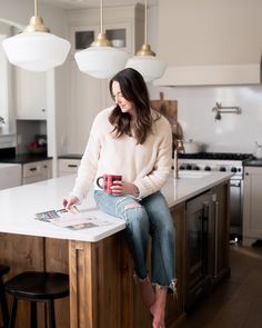 a woman sitting on top of a kitchen counter holding a coffee cup and reading a magazine