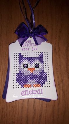 a cross stitch owl ornament hanging on a wooden table with purple ribbon and bow