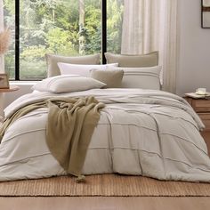 a bed with white sheets and pillows in front of a large window that has trees outside