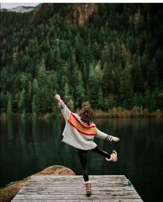 a woman standing on top of a wooden dock next to a body of water with trees in the background