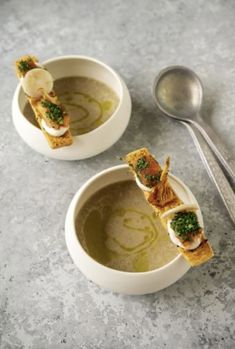 two white bowls filled with soup and garnished with green toppings next to a spoon