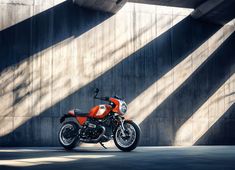 an orange motorcycle parked in front of a concrete building with long shadows on the wall