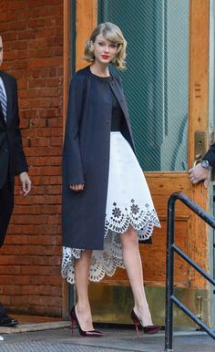 a woman in a black coat and white dress walking out of a building with her hand on her hip