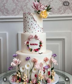 a three tiered wedding cake with flowers on the top and bottom, sitting on a glass table in front of a wall