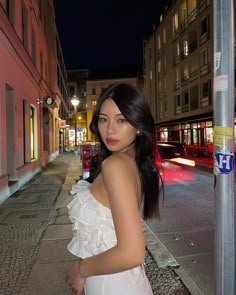 a beautiful young woman standing next to a pole on a city street in front of buildings