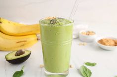 a green smoothie in a tall glass next to some bananas and avocado