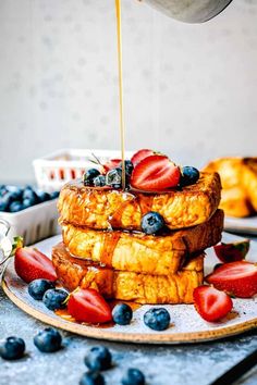 french toast with syrup being drizzled over it and topped with strawberries and blueberries