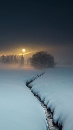 the sun is setting over a snowy field with water running through it and trees in the distance