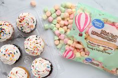 cupcakes with frosting and sprinkles in a plastic container next to a bag of marshmallows