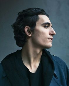 a man with dark hair and black shirt looks off to the side while standing in front of a gray wall