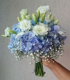 a bouquet of blue and white flowers in someone's hand with a gray wall behind it